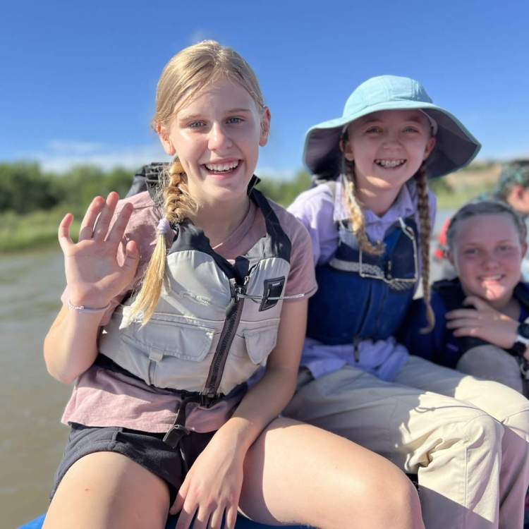 3 smiling girls in life jackets sit on a raft floating down a river