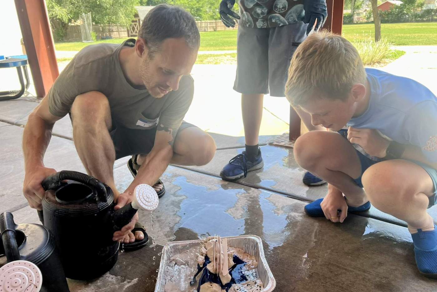 boy watches as man pours water from watering can onto her clay dam model in metal tray