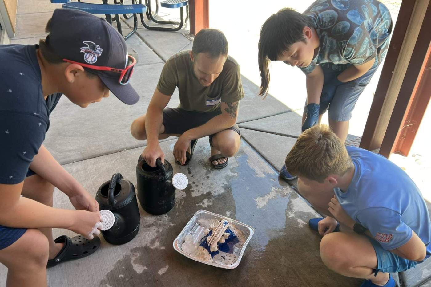 3 boys around metal tray holing clay dam model while man pours water from watering can