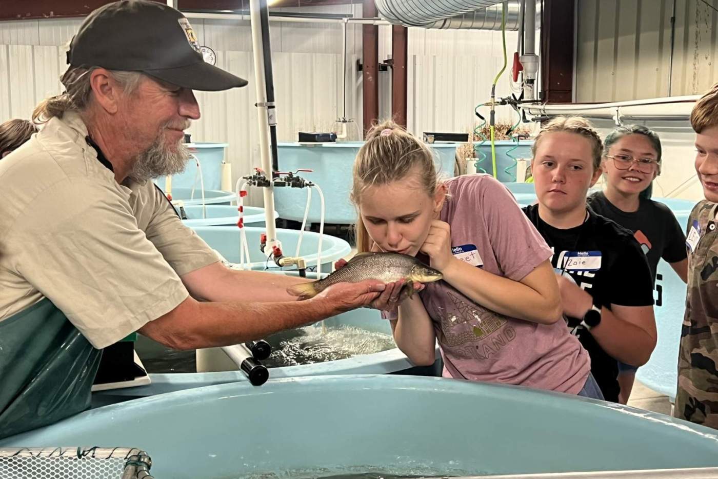 girl kisses a large fish held by man in a hat