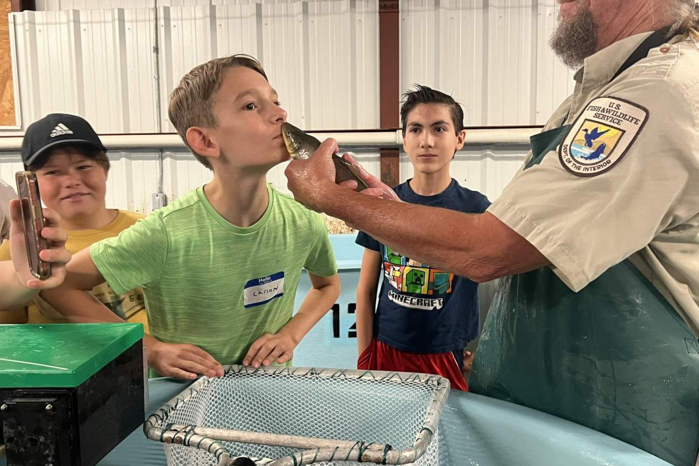 boy kisses a fish held by man in a hat