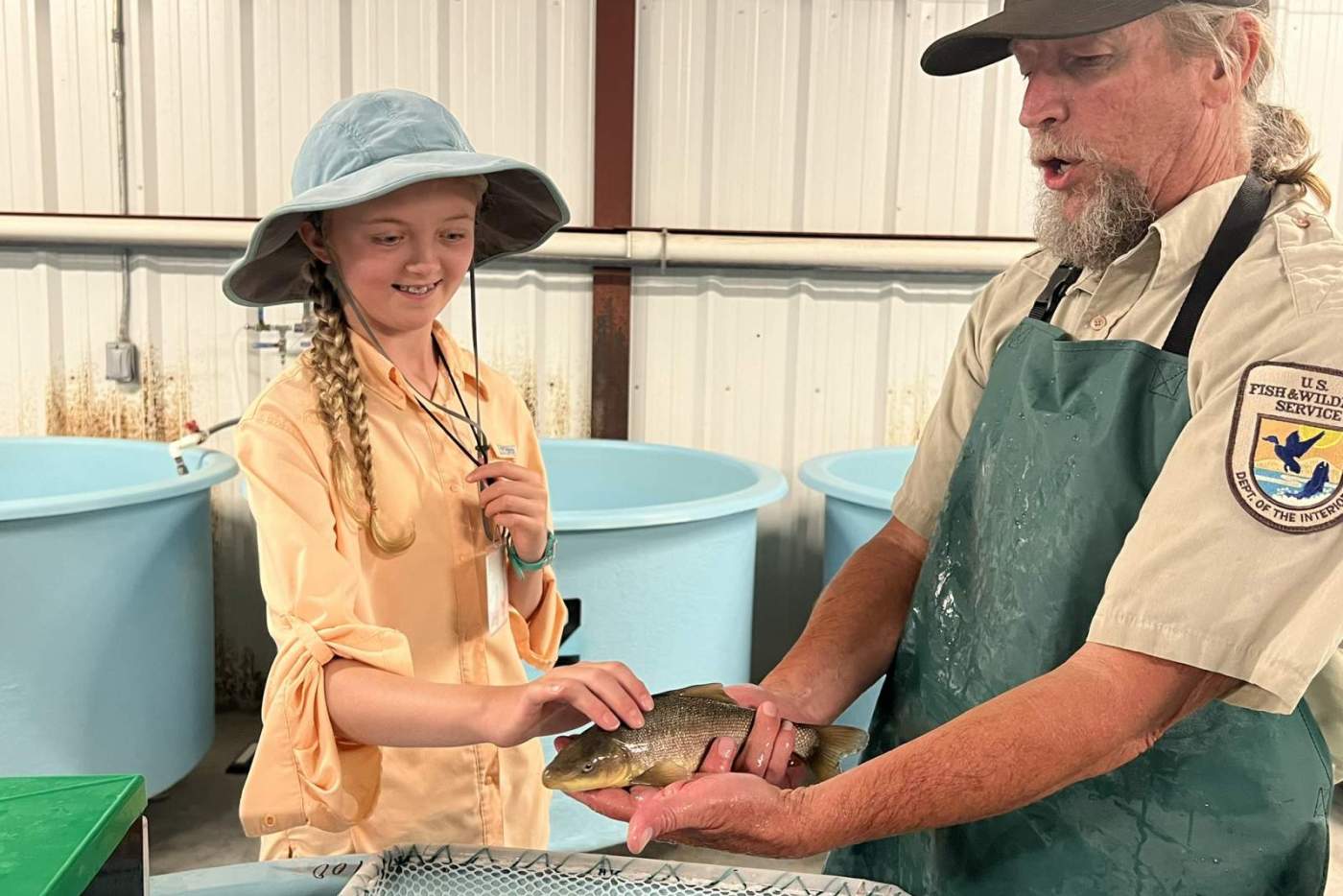 girl touches a fish held by man in a hat and apron