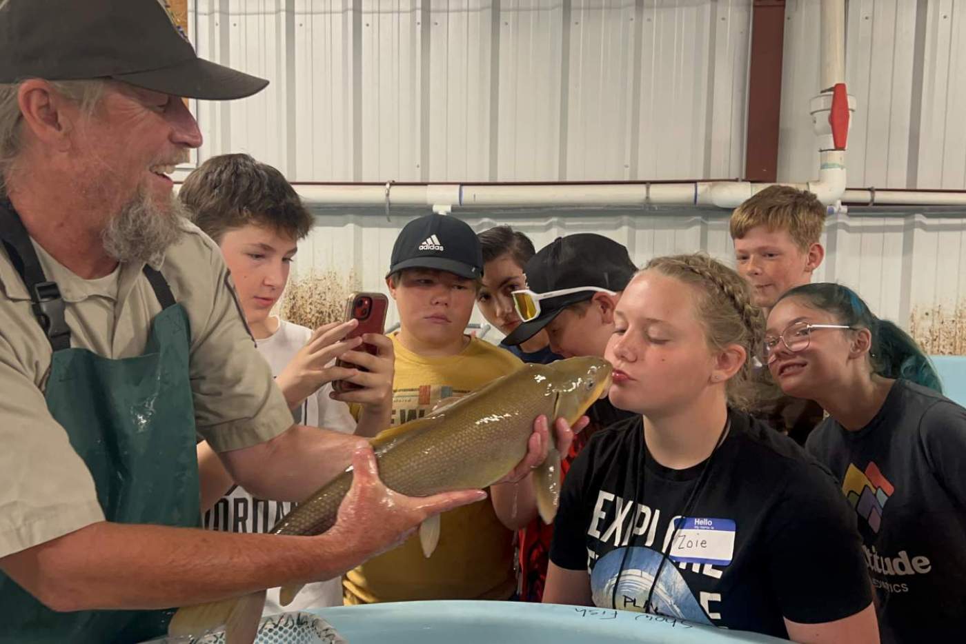 girl kisses a large fish held by man in a hat