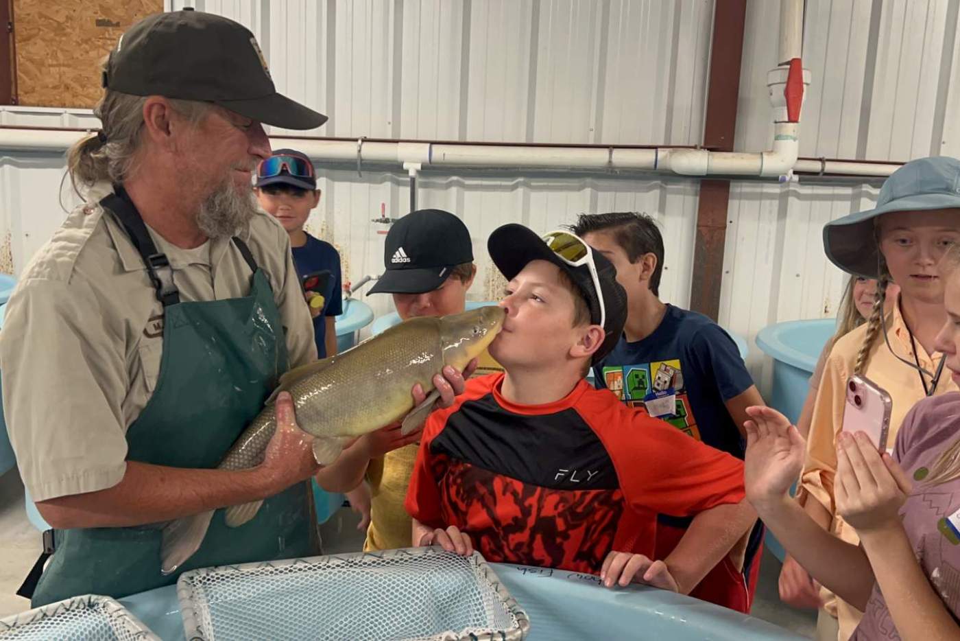 boy kisses a large fish held by man in a hat, while other children look on