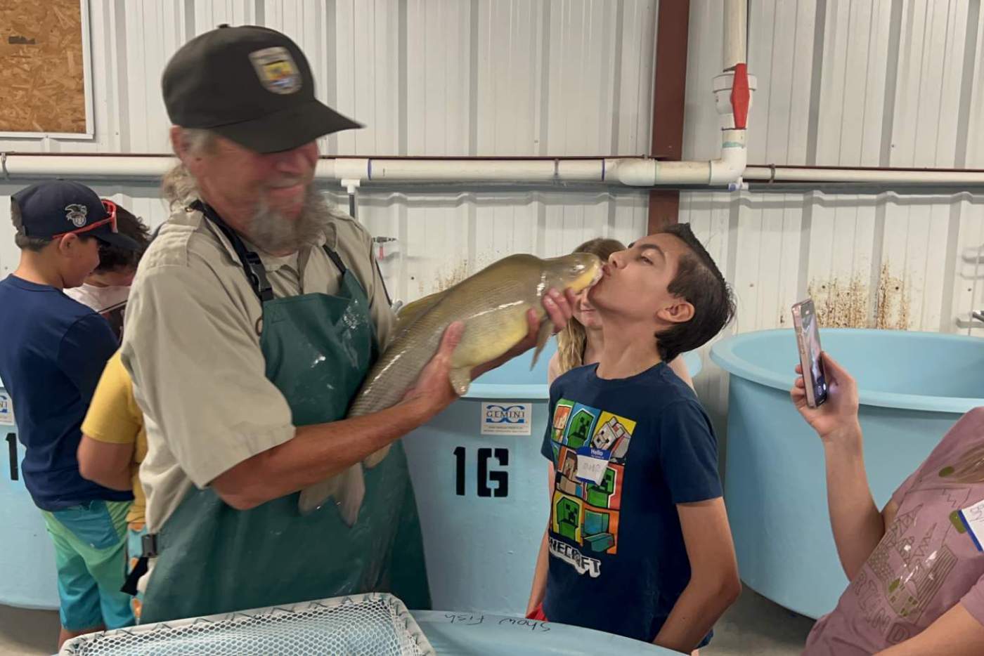 boy kisses a large fish held by man in a hat