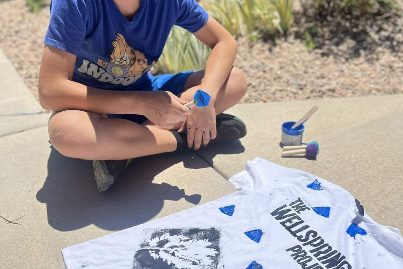 Boy sits on the ground with the shirt he designed that says "The Wellspring Project"