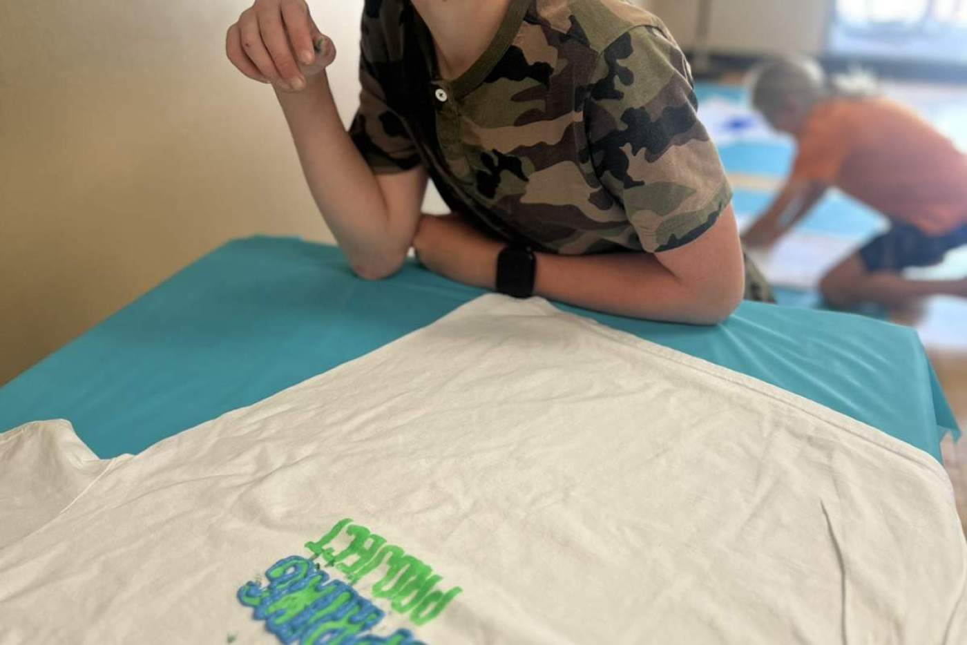 Boy lays on the floor with the shirt he designed that says "The Wellspring Project"