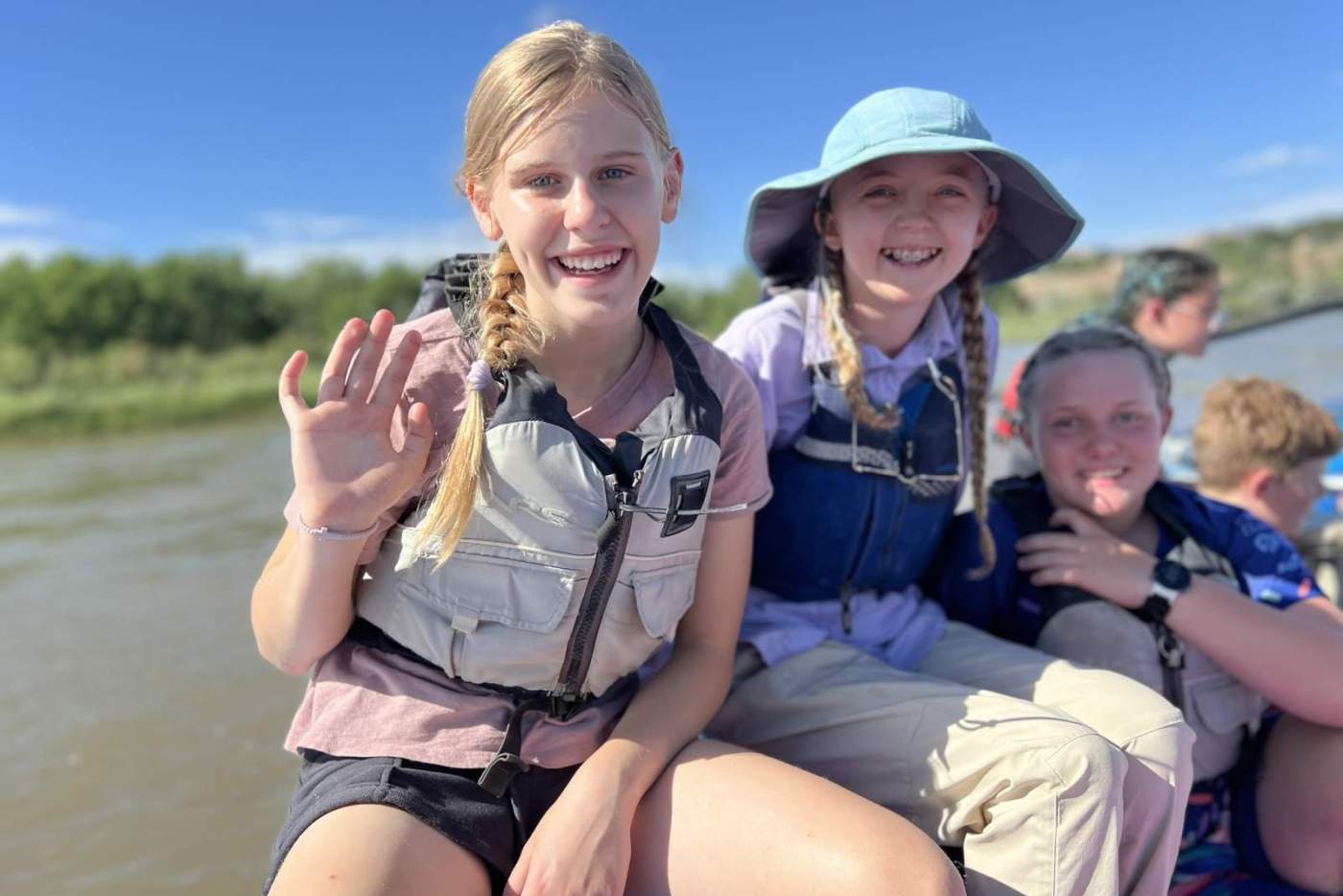 3 smiling girls in life jackets sit on a raft floating down a river