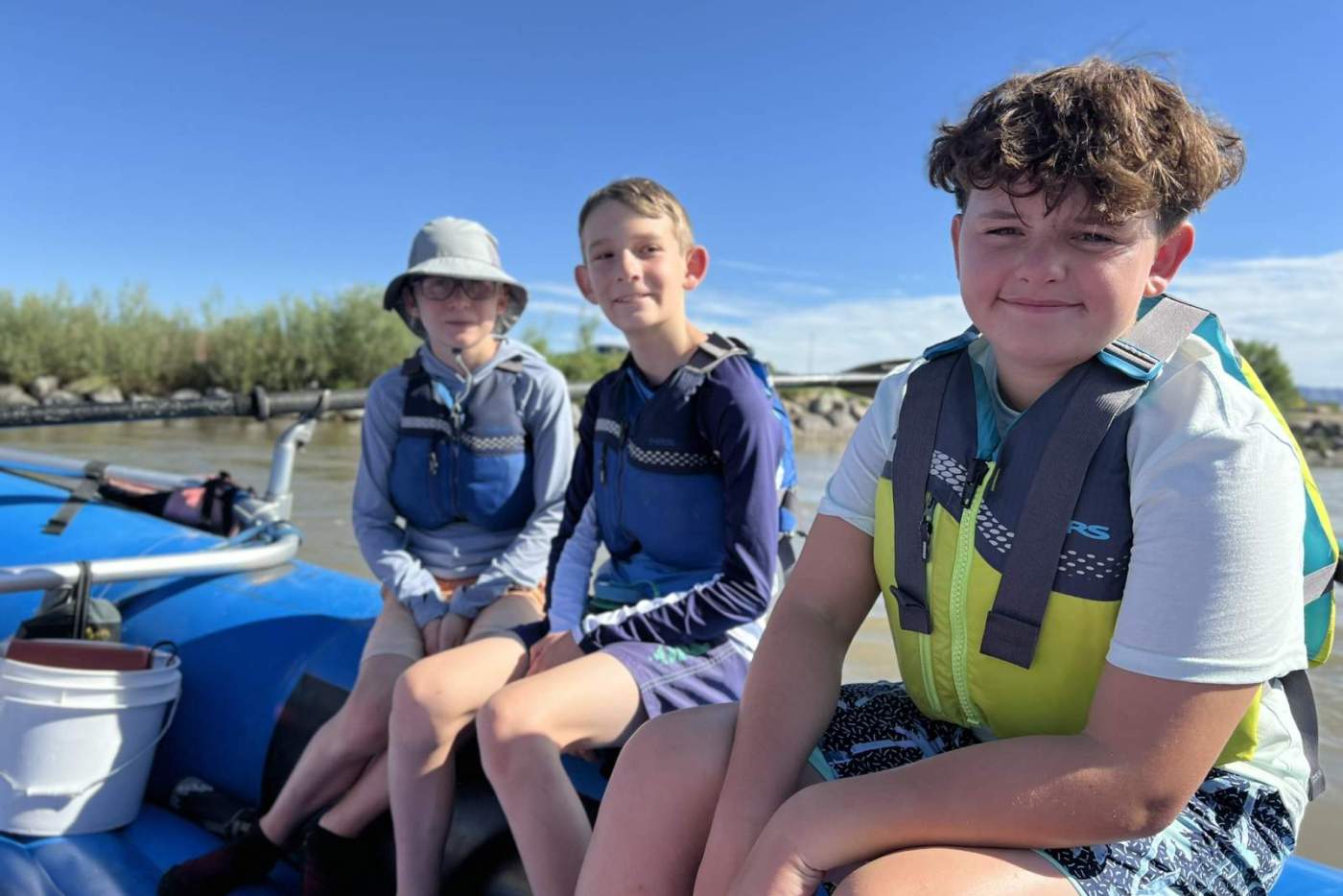 2 boys in life vests sitting on a blue raft on the river