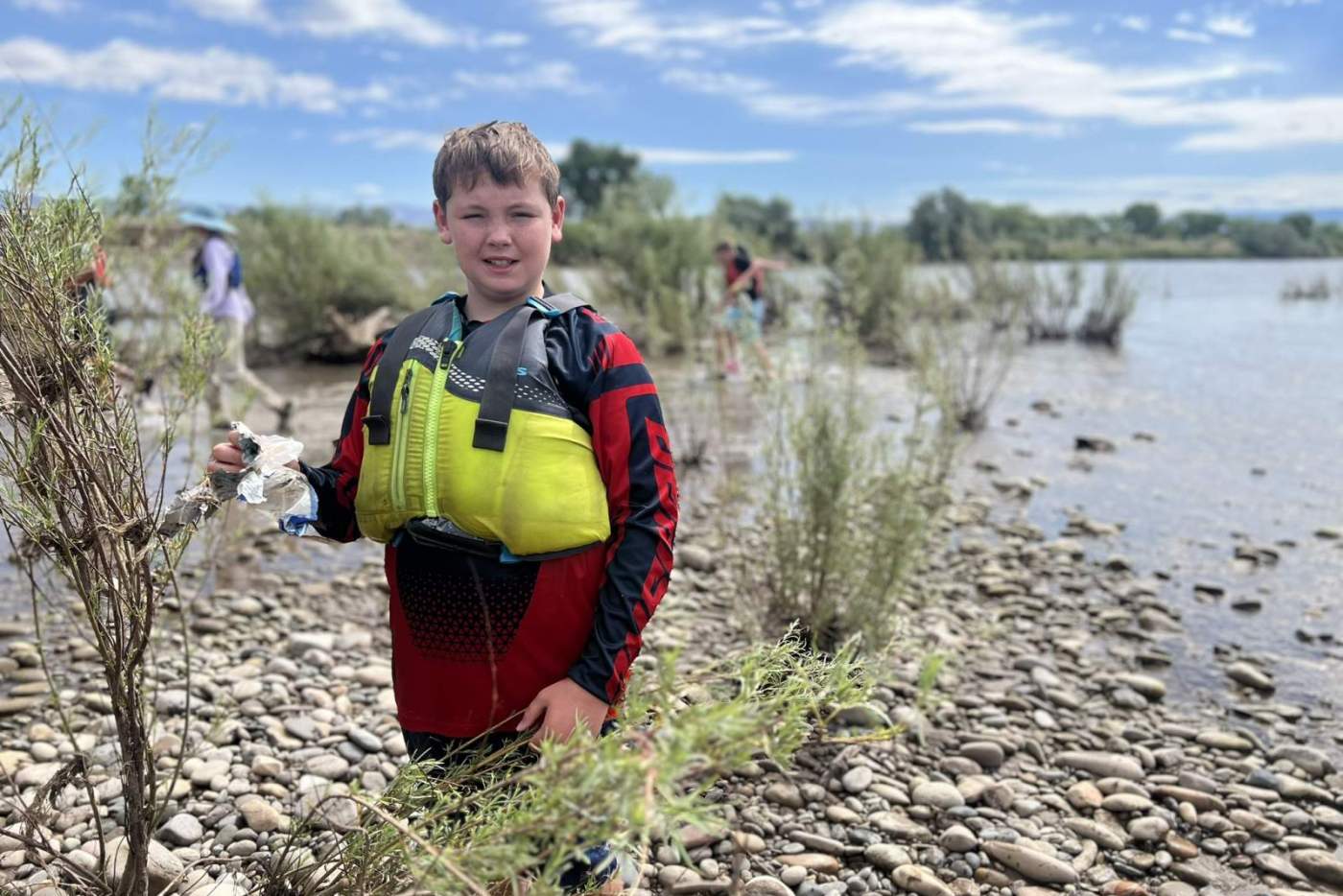 Boy in red shirt with yellow life vest standing on rocky riverside with plants