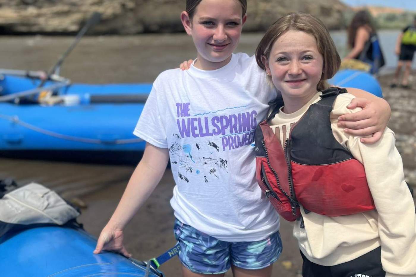 2 girls, 1 wearing a life vest, with an arm around eachother, stand next to a blue raft in the river