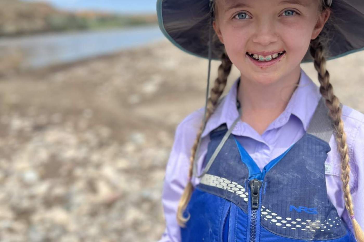 a girl in a blue sunshirt, blue sun hat, and blue life vest, holds object and smiles while standing on the riverside