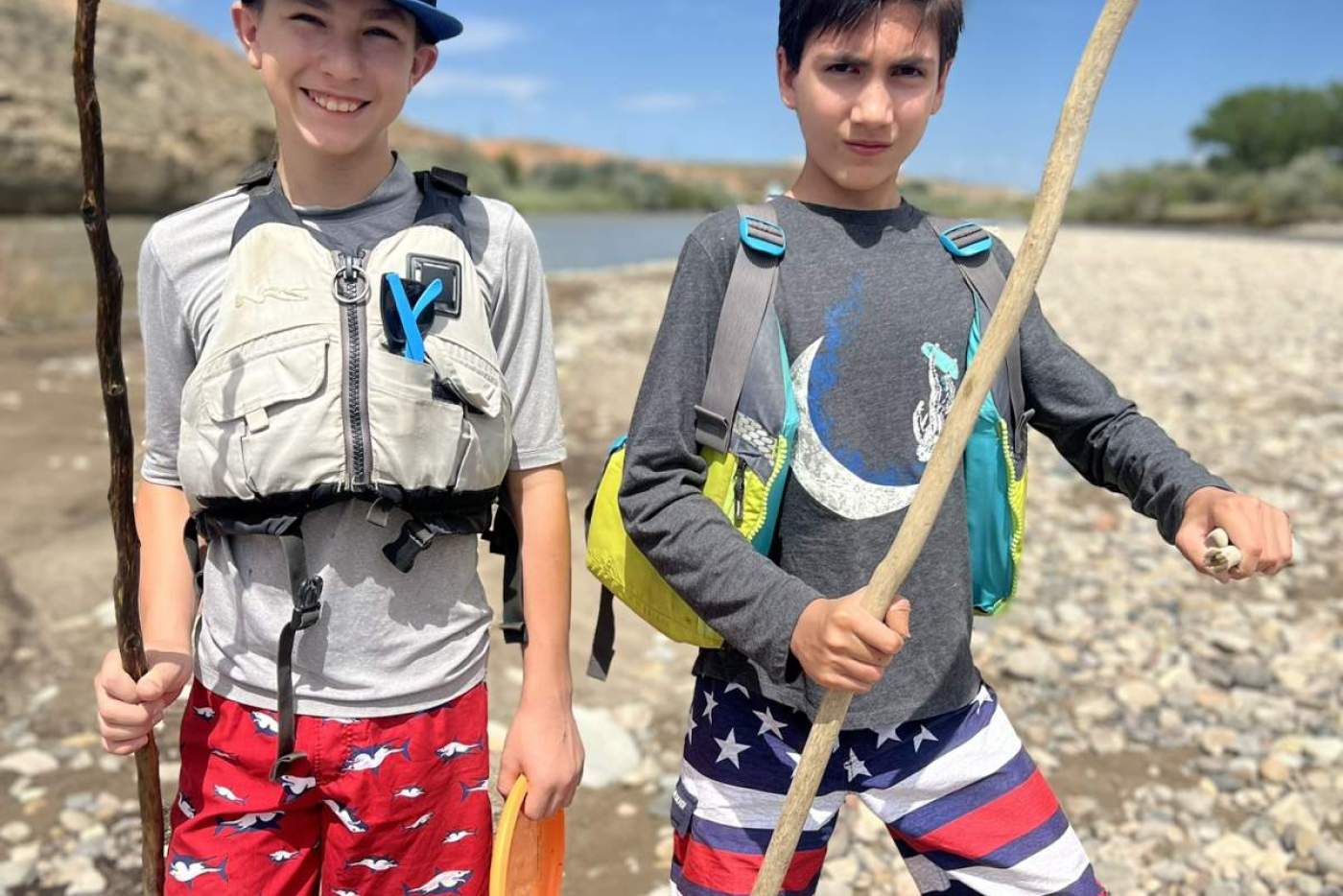 2 boys in life vests pose on a rocky riverside holding sticks