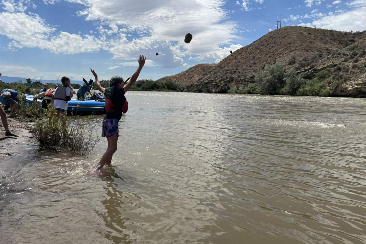 young person in life vest throws a rock into the river with blue raft and mountains in the backgrouns