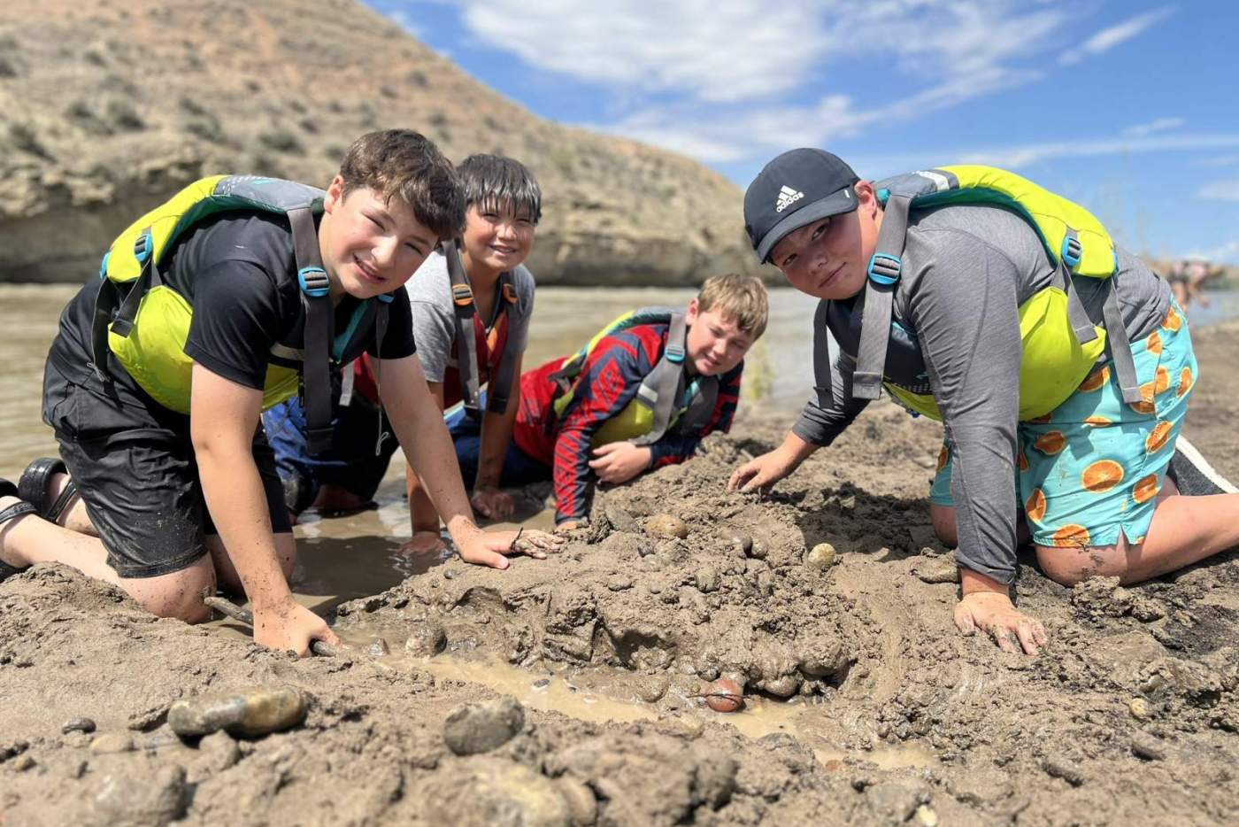 4 boys in life vests building structures in the sand along the riverside