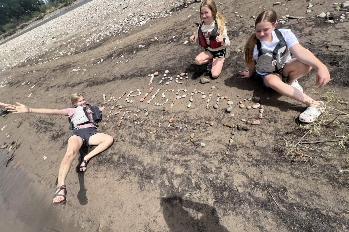 3 girls posing on the ground next to "The Wellspring Project" spelled in river rocks