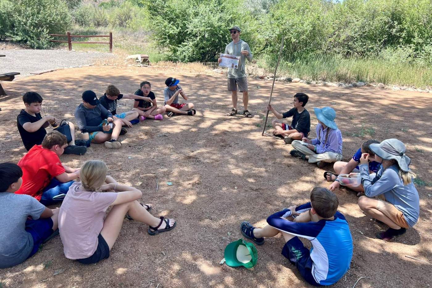 group of middle schoolers sitting in the shade under a tree listening to a standing man